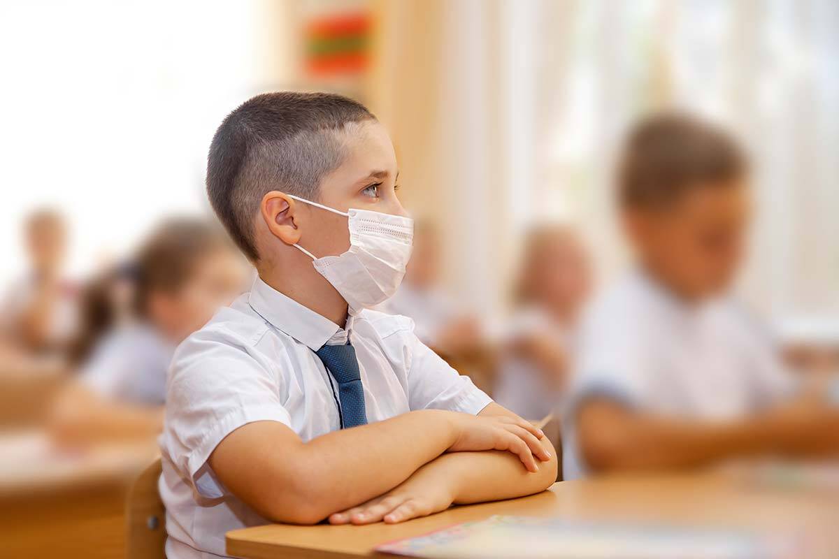 Boy in school uniform wearing a face mask at a desk, he is surrounded by other children in masks.